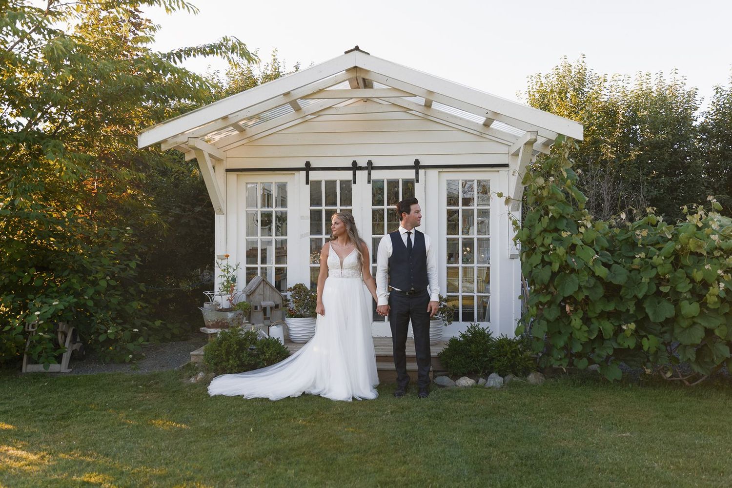 Bride and groom stand together in front of a charming white garden shed surrounded by lush greenery.