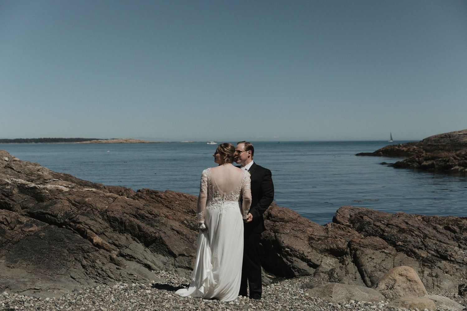 Bride and groom standing on rocky coastal shoreline with stunning blue ocean and clear sky behind them.