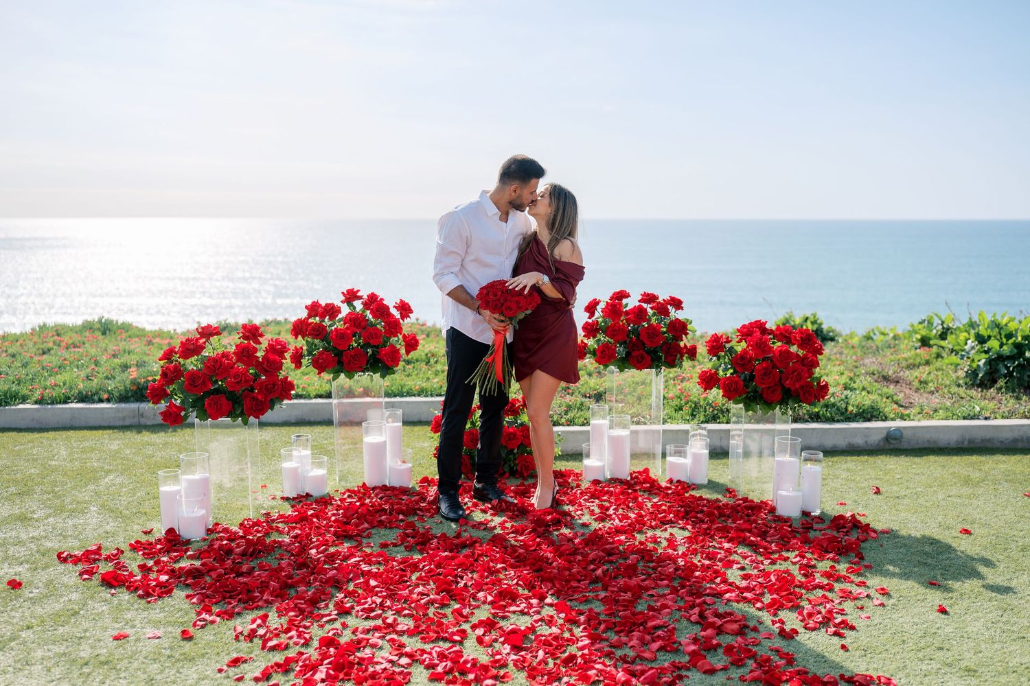 Red rose petals scatter across a grassy lawn as figures in formal attire walk through the romantic scene.
