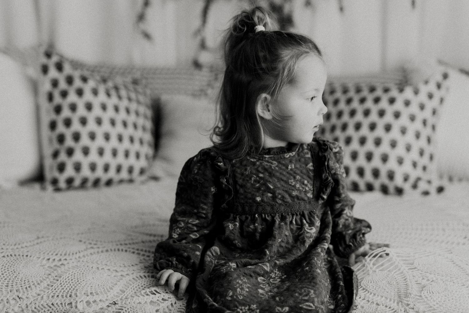 A black and white photo series shows a young child in a dark dress playing on a bed with polka dot pillows.