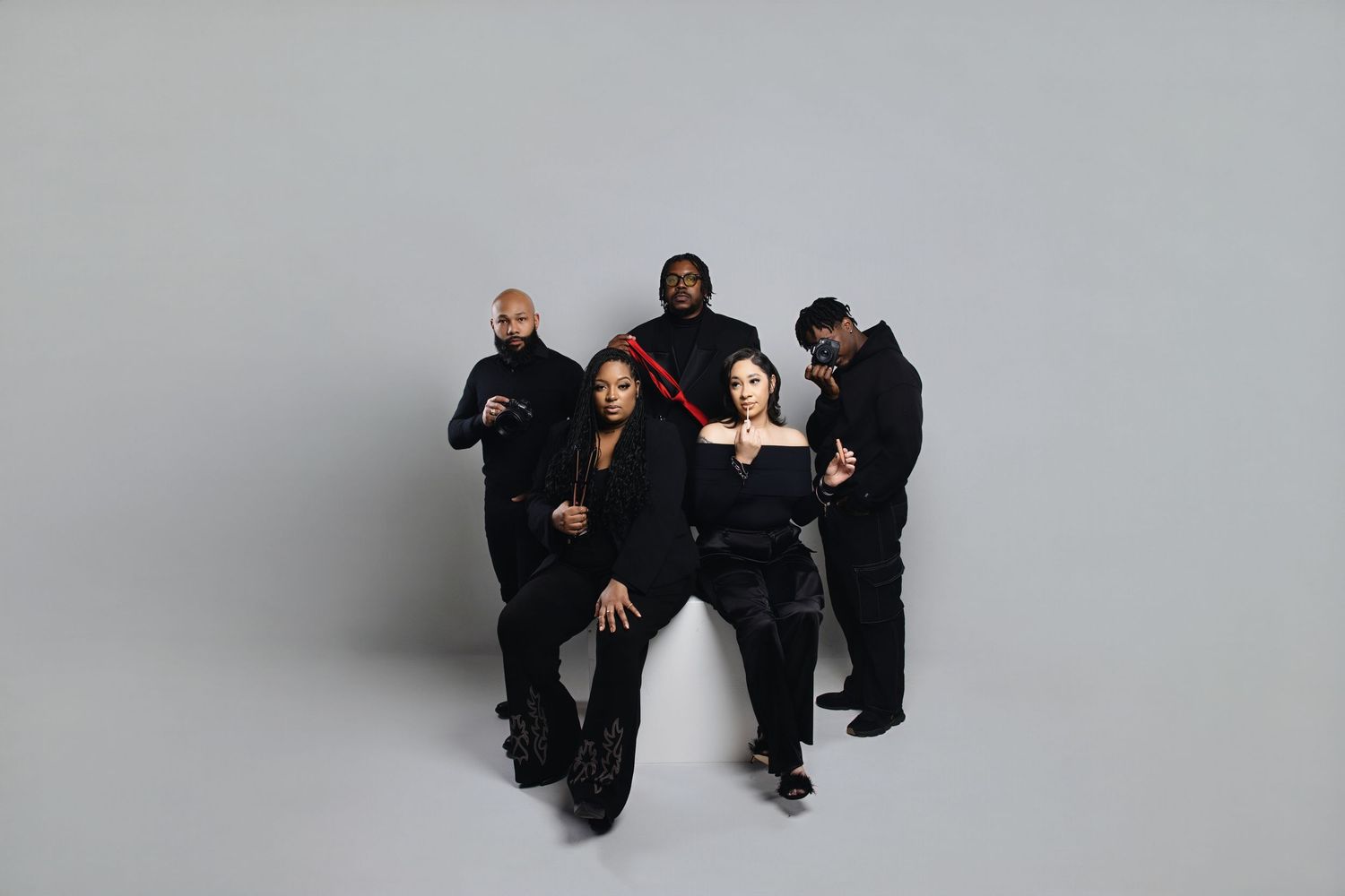 Three people in black outfits pose together in various casual positions against a gray backdrop.