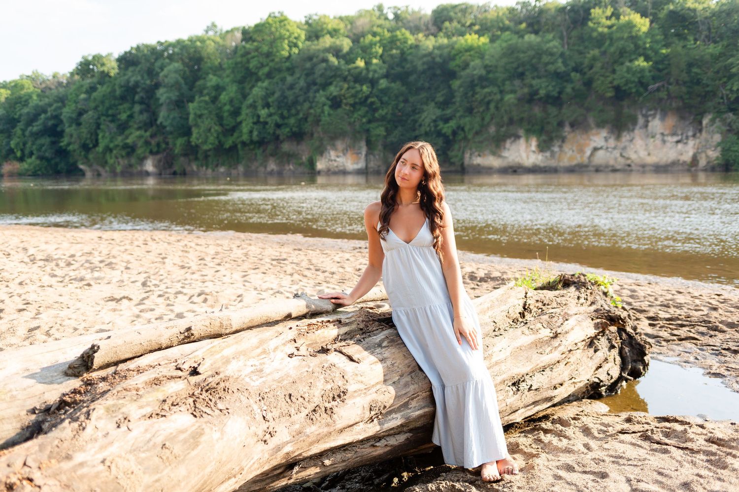 A person in a flowing white dress poses on driftwood at a scenic riverside beach with cliffs and trees in the background.