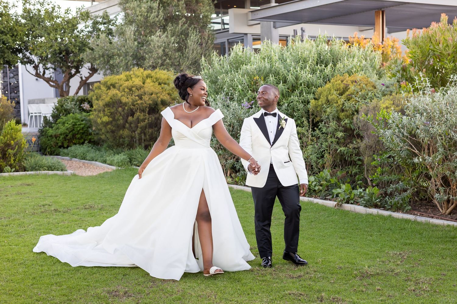 A couple in wedding attire pose together on a green lawn with lush garden foliage in the background.