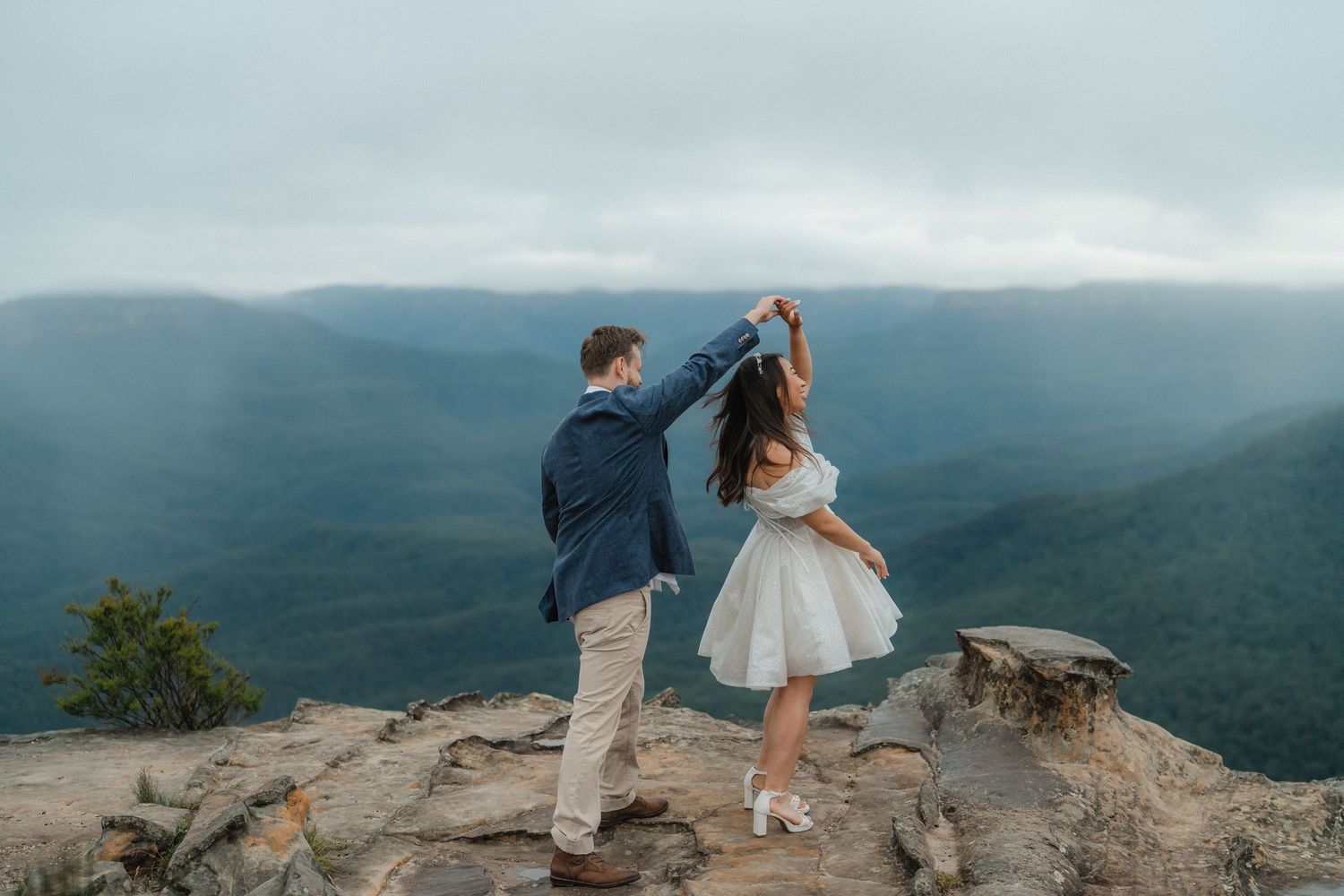 A couple shares a tender moment together on a scenic mountain overlook with dramatic mountain ranges in the background.