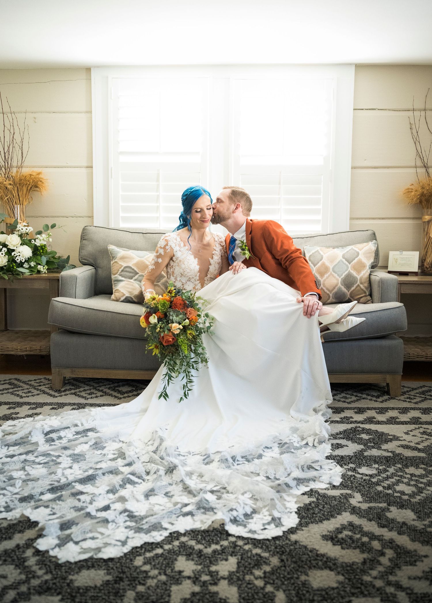 A couple shares a romantic moment on a gray couch, with the bride wearing a lace gown and holding a vibrant fall bouquet.