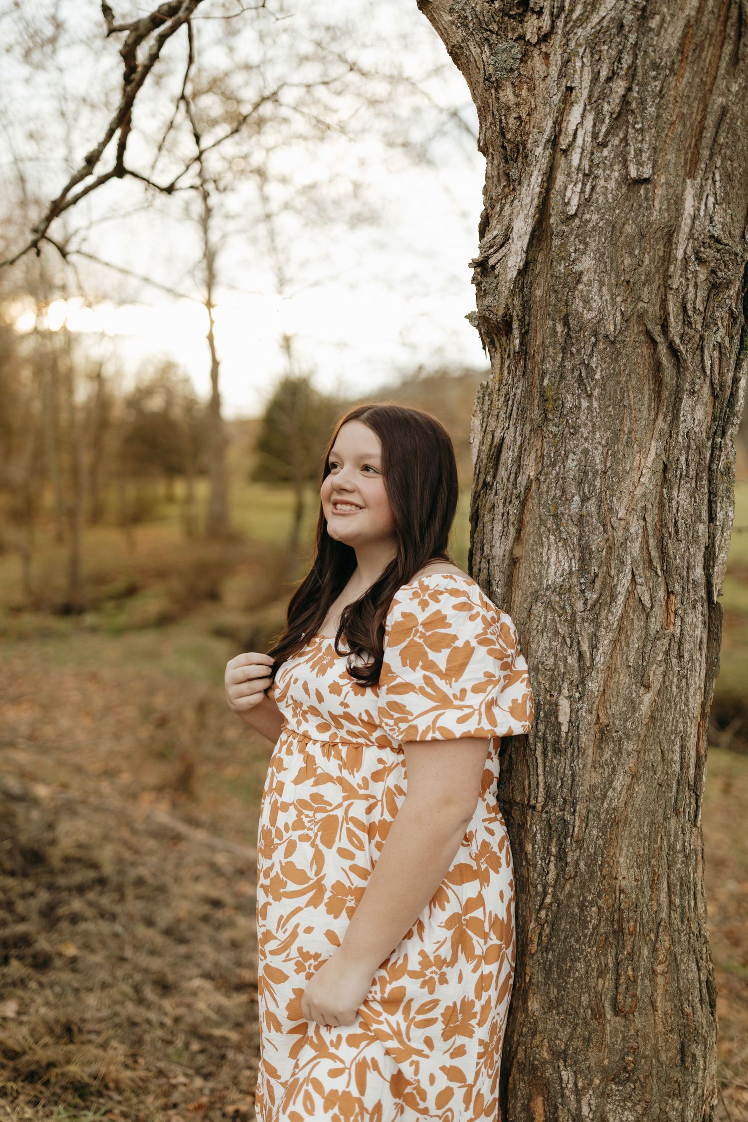 A person in a floral dress leans against a large tree trunk during golden hour in an autumn setting.