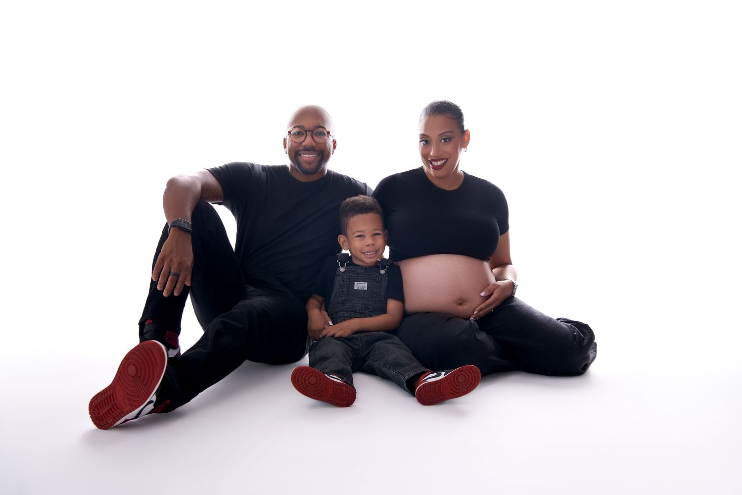 Three people wearing black clothing and red shoes lie on a white background in a playful pose.