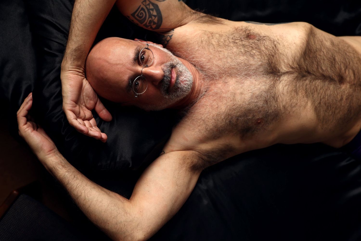 Male boudoir client laying on black silk sheets in a Seattle Airbnb, shirtless, looking at camera.