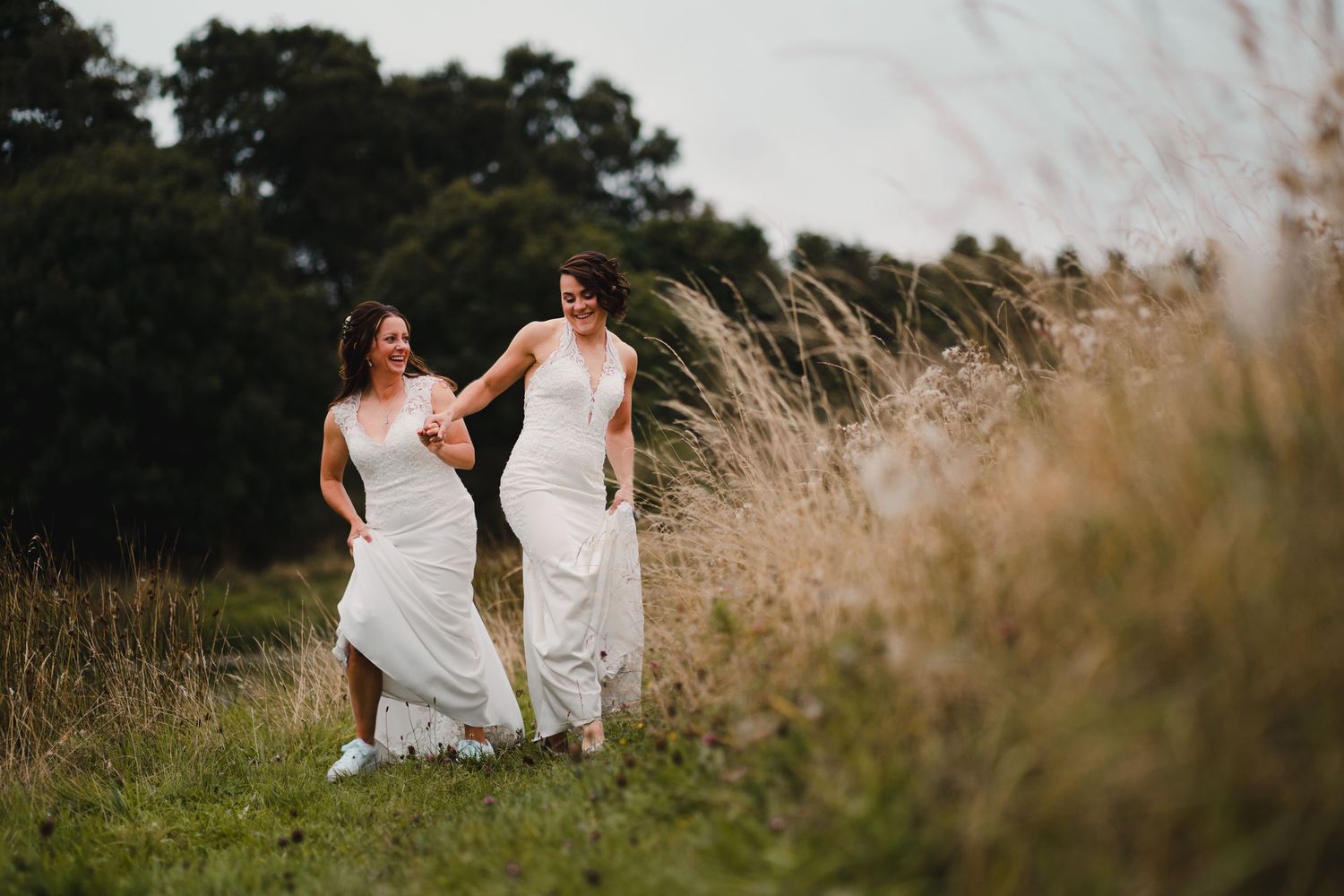 Two people in long white dresses walk together through a serene field of tall grass at sunset.