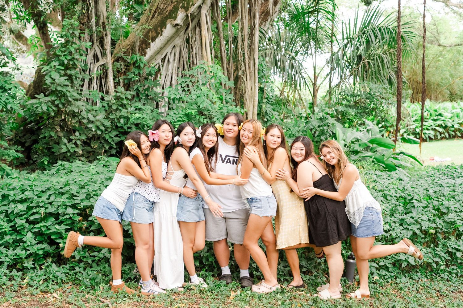 A group sits together outdoors on tree roots surrounded by lush green foliage in a natural setting.