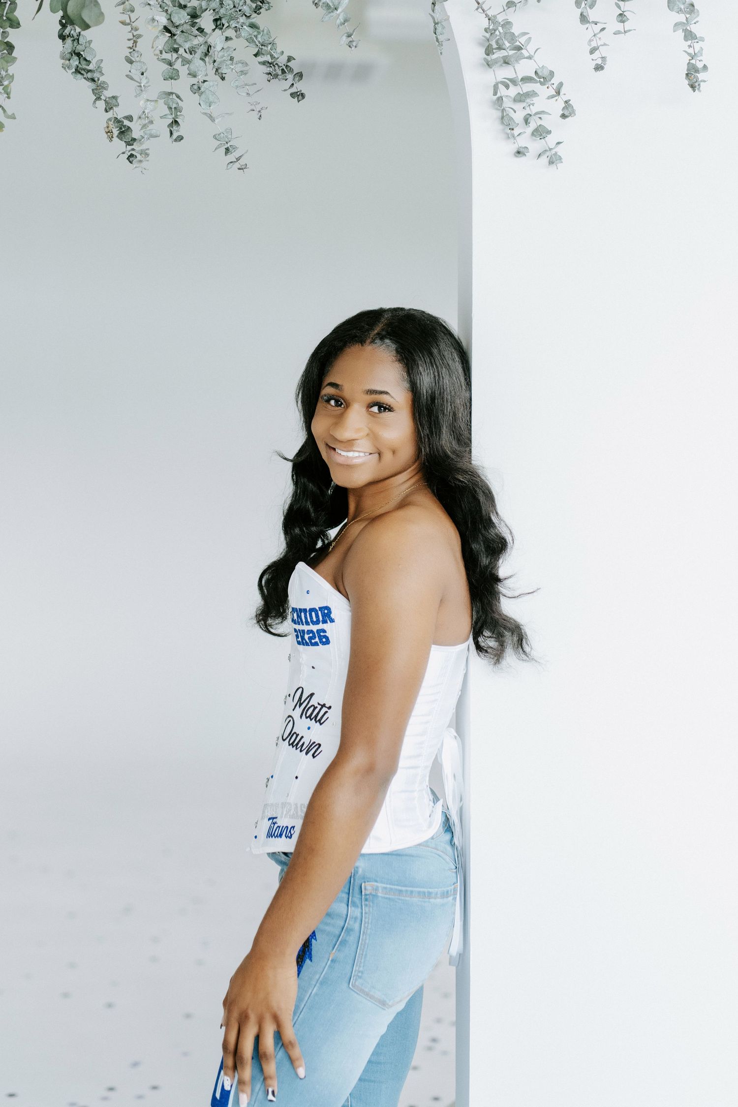A person in a white tank top and light blue jeans poses playfully against a light gray background.