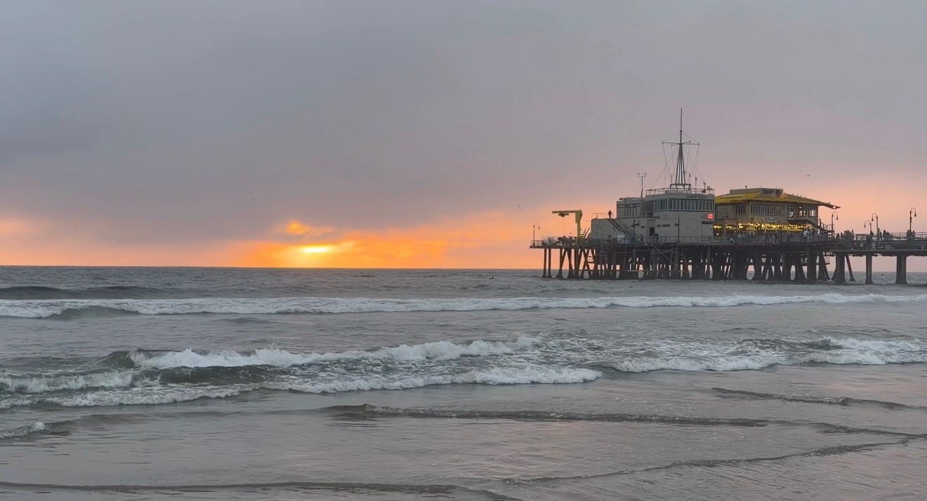 Panoramic view of a beach pier at sunset with crashing waves and orange sky over the ocean horizon.