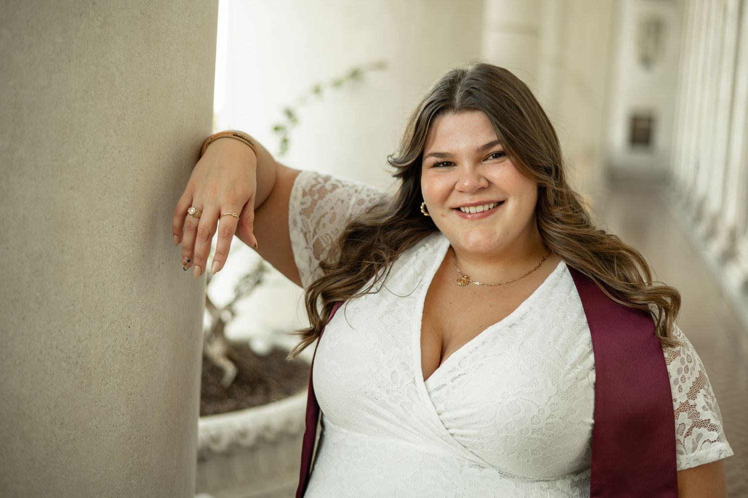 Plus size model in white lace dress and burgundy vest poses against a light colored wall with a bright smile.