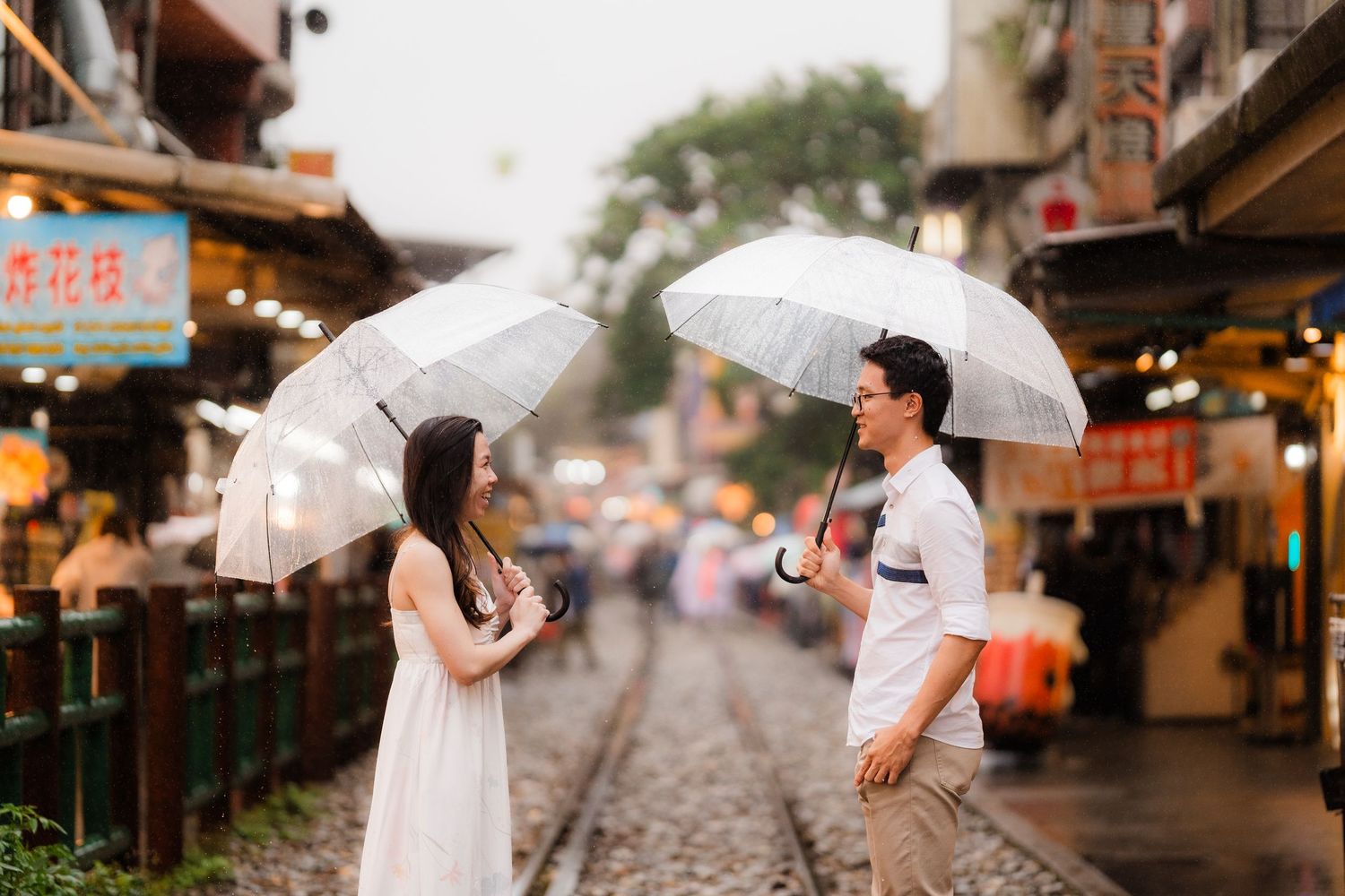 Couple with white umbrellas walking along a rainy street lined with traditional Asian storefronts and lanterns.