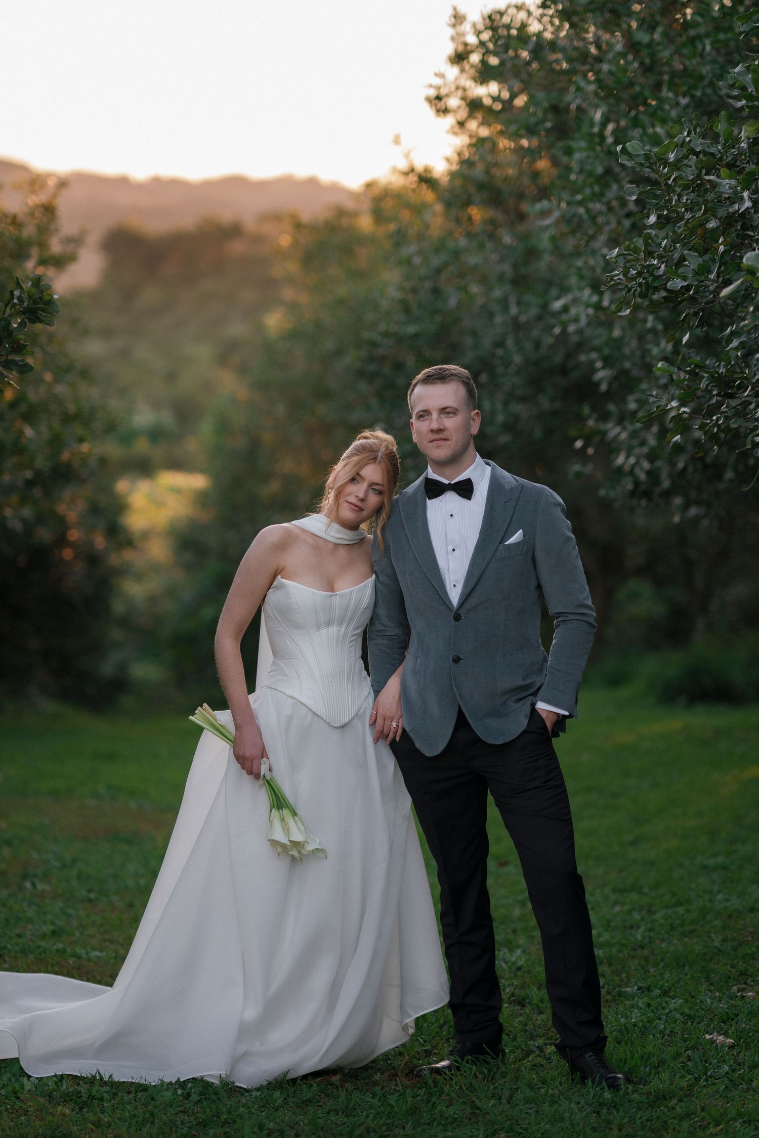 A couple in wedding attire poses together in a lush garden setting during golden hour.