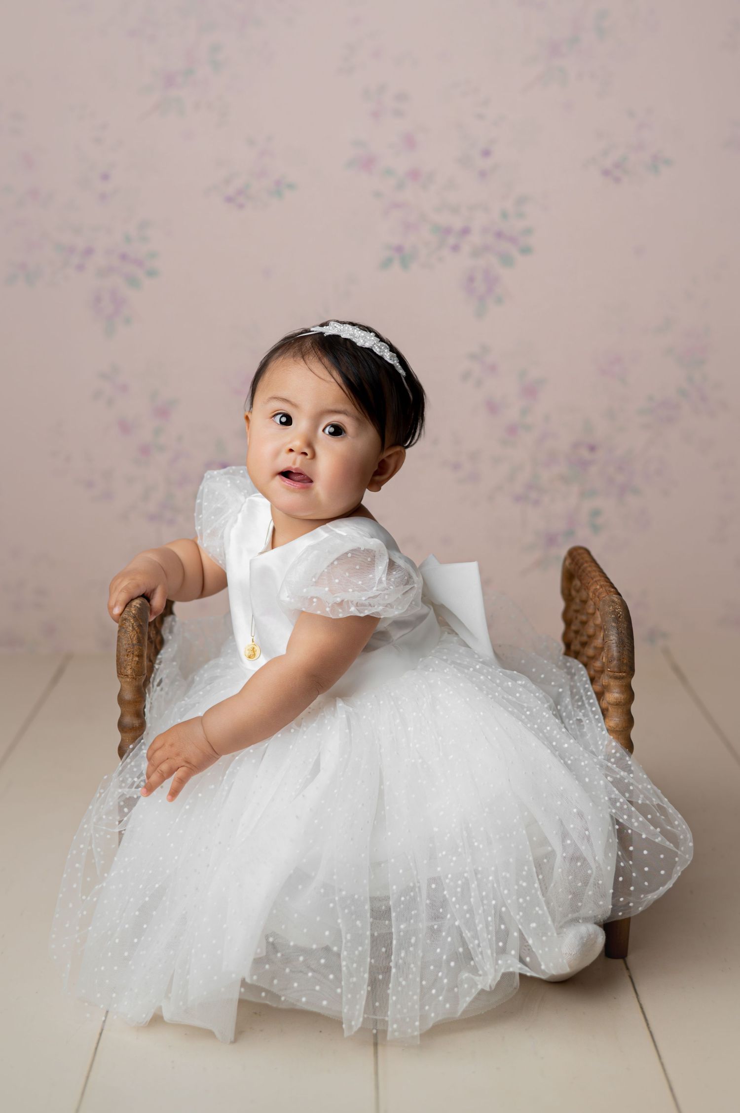Sweet toddler in pink tulle dress and boots sits on vintage trunk against floral wallpaper background.