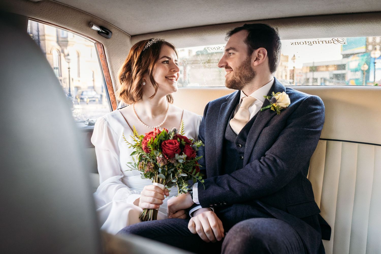 A newly married couple sits in the back of their wedding car, looking into each other's eyes.