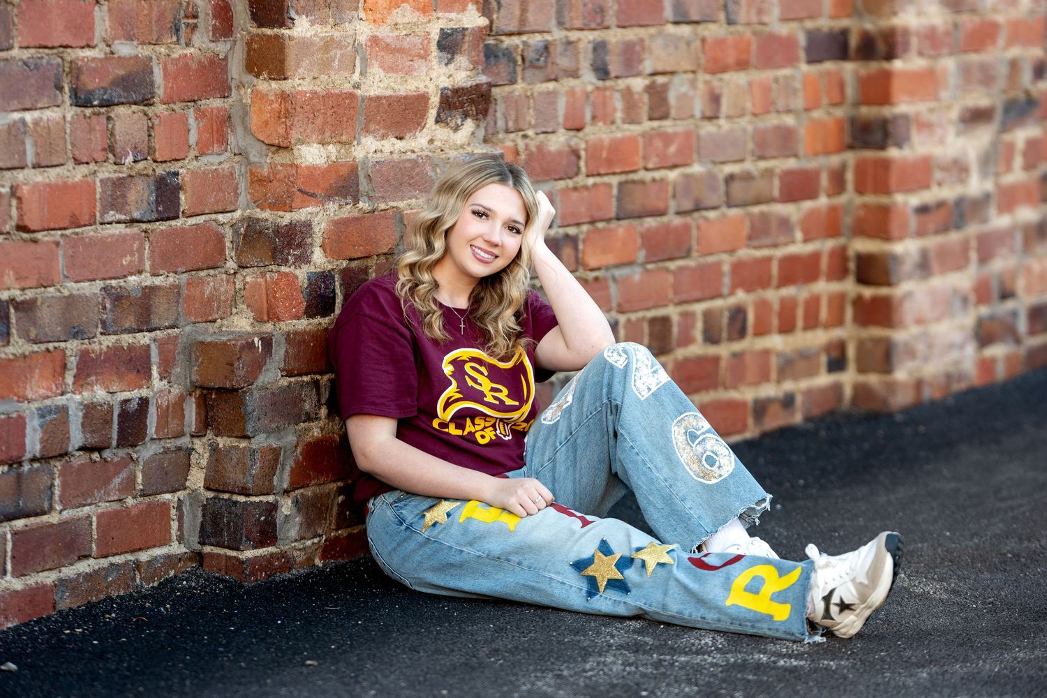 Blonde teen girl in maroon shirt and painted jeans sitting against brick wall.