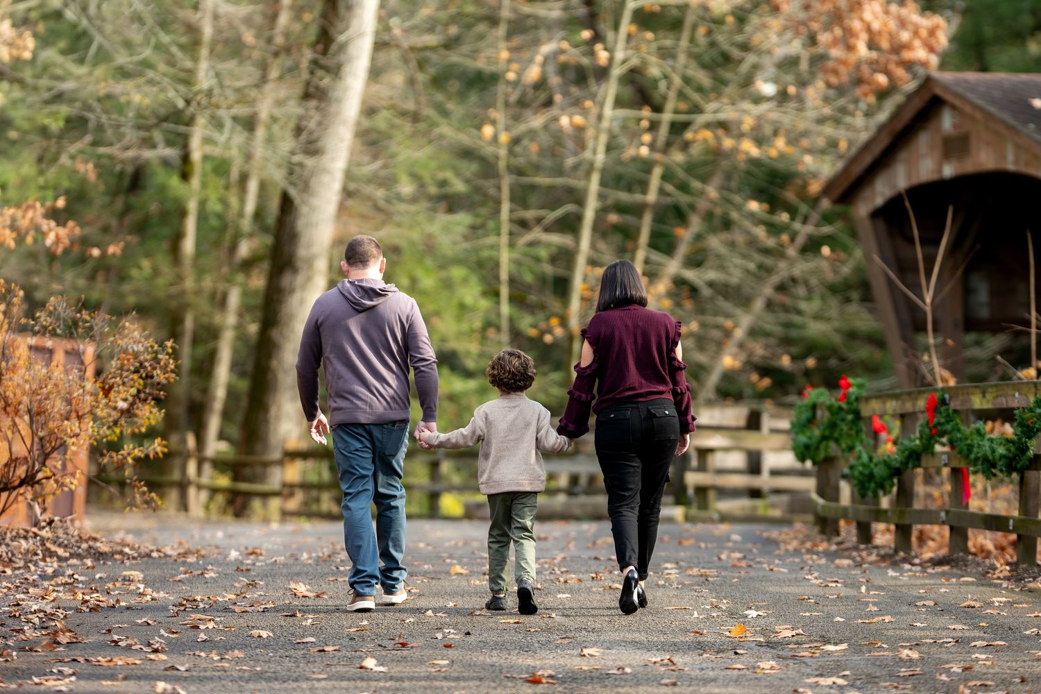 Family of three walks hand-in-hand down a path near a covered bridge decorated with Christmas wreaths.