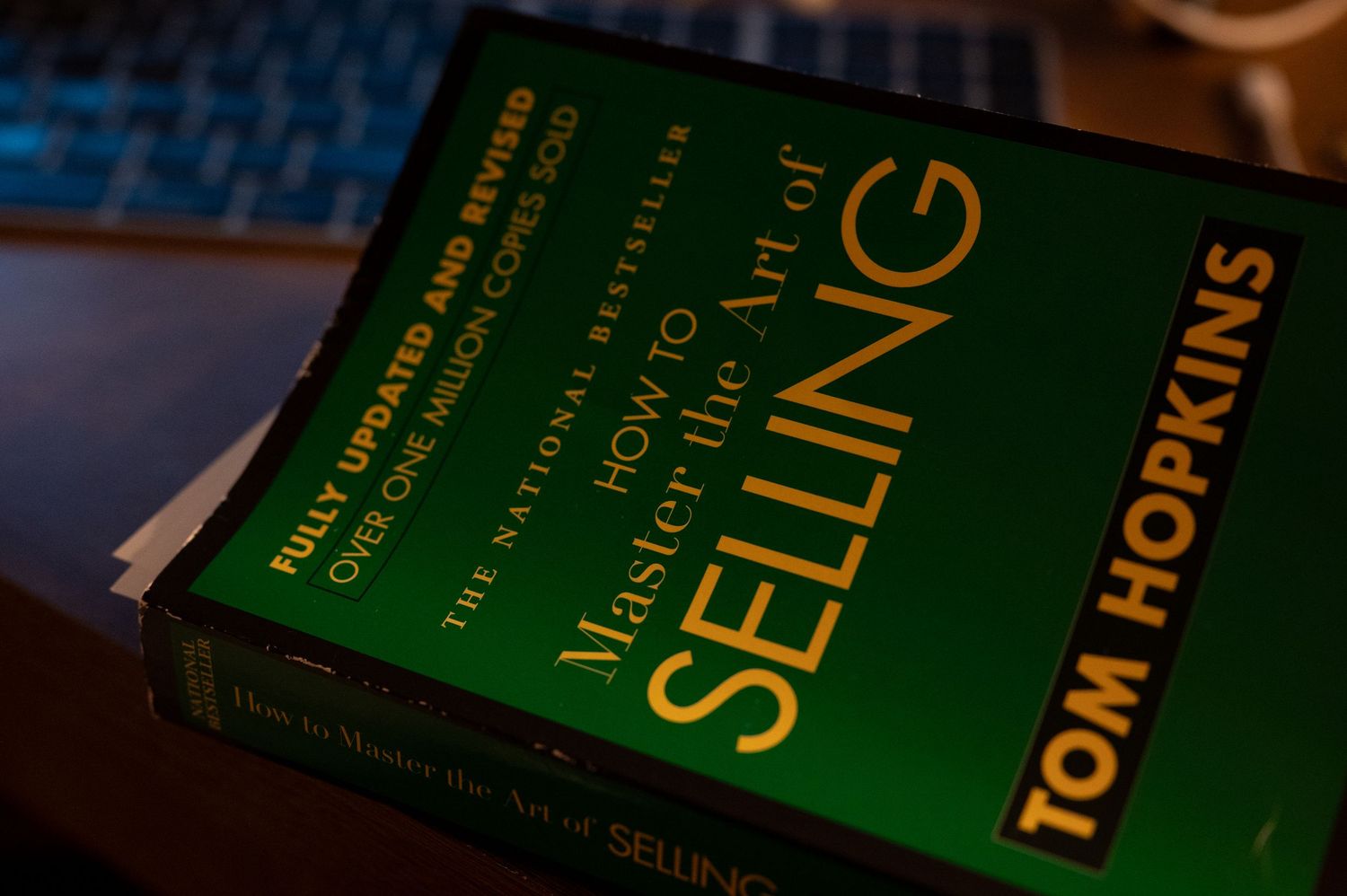 A green hardcover book titled 'The Art of Selling' by Tom Hopkins sits on a dark surface.