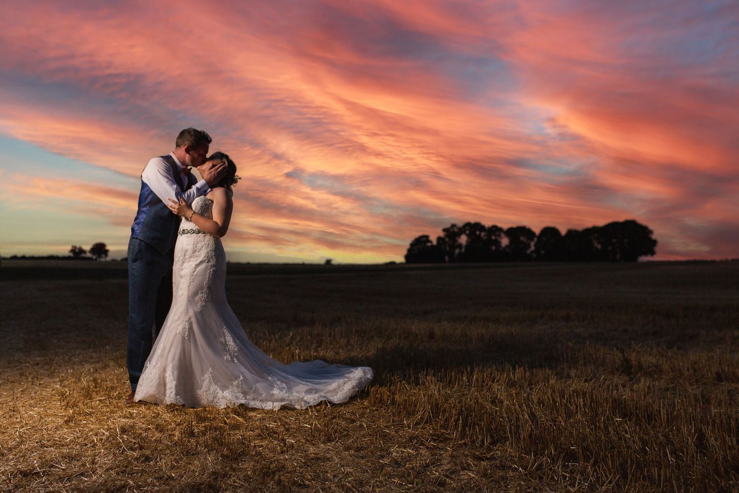 A couple in wedding attire embraces against a dramatic orange and pink sunset sky over a rural field.