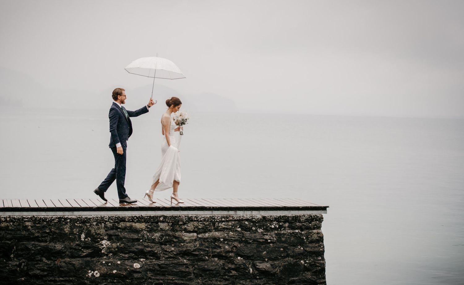 Elegant bride and groom walking on stone wall with white umbrella against misty lake.