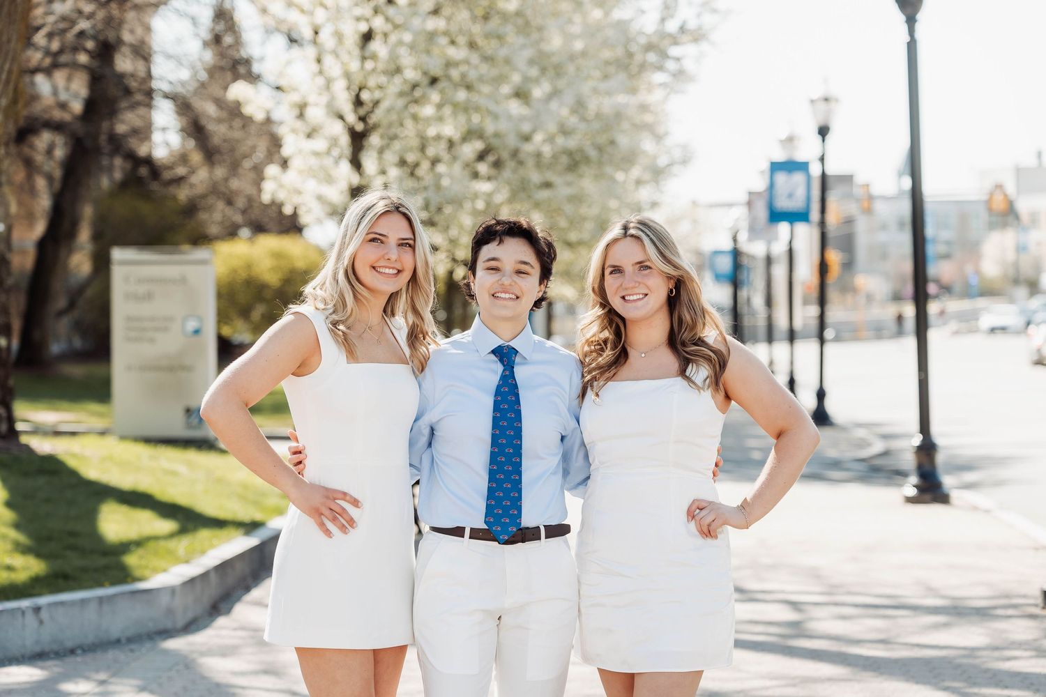 Three people in white outfits pose together on sunny sidewalk with street lamps.