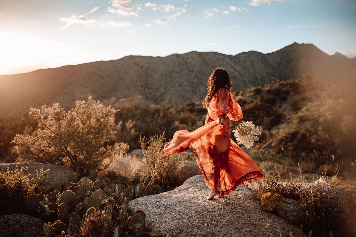 A flowing red dress catches the wind against a mountainous desert landscape at golden hour.