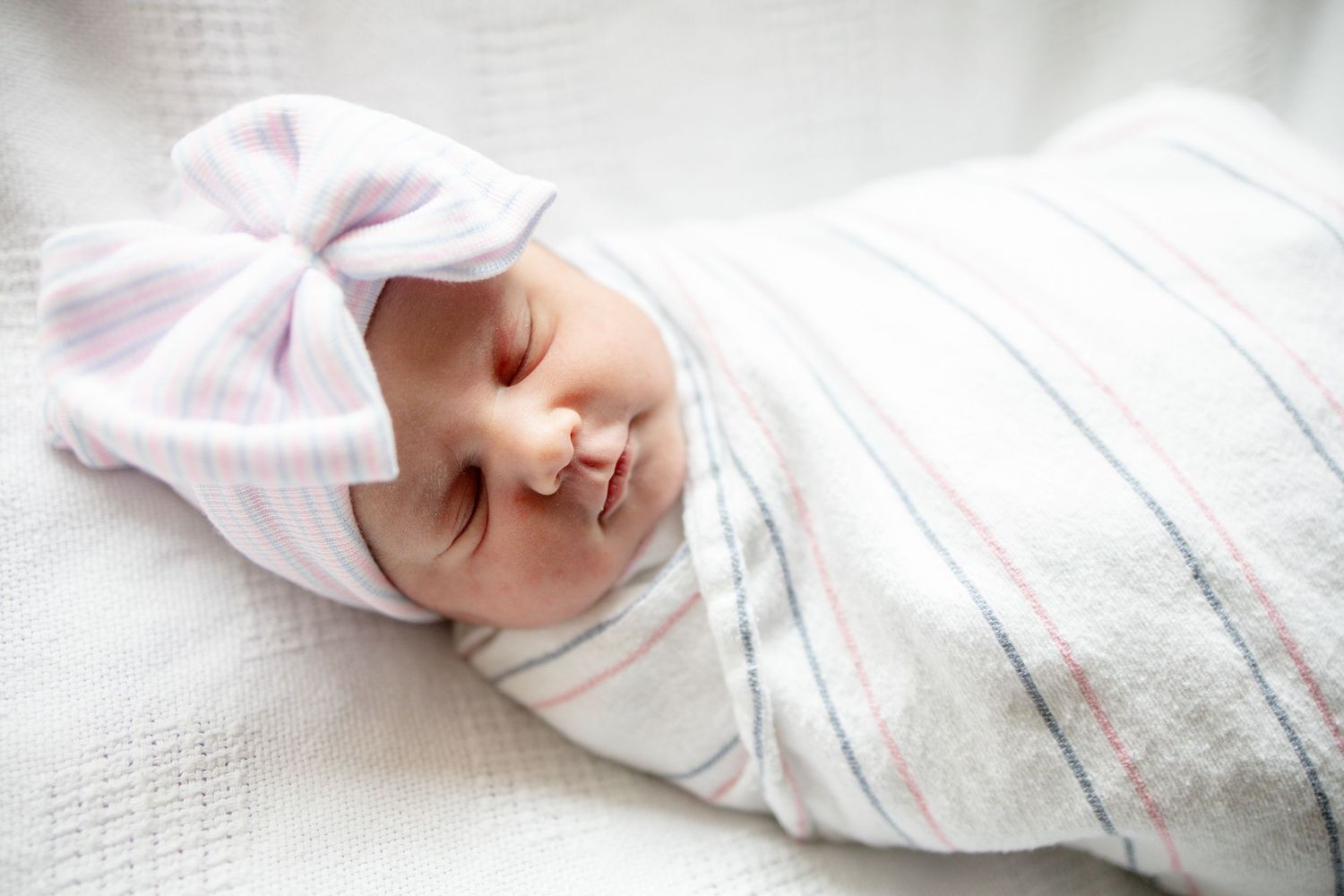 Newborn baby swaddled in white striped blanket wearing a white bow hat, sleeping peacefully.