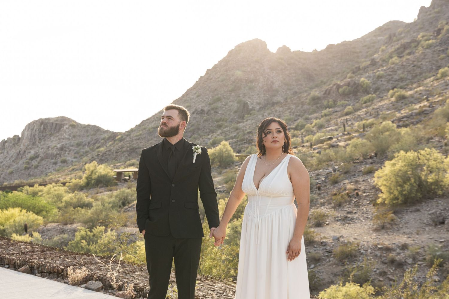 A couple in wedding attire stands together against a scenic desert mountain backdrop at sunset.