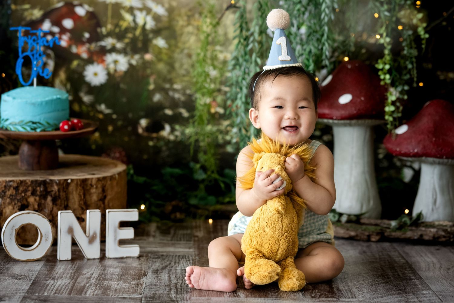 First birthday celebration setup with decorative mushrooms, cake, and a joyful baby holding a yellow teddy bear.