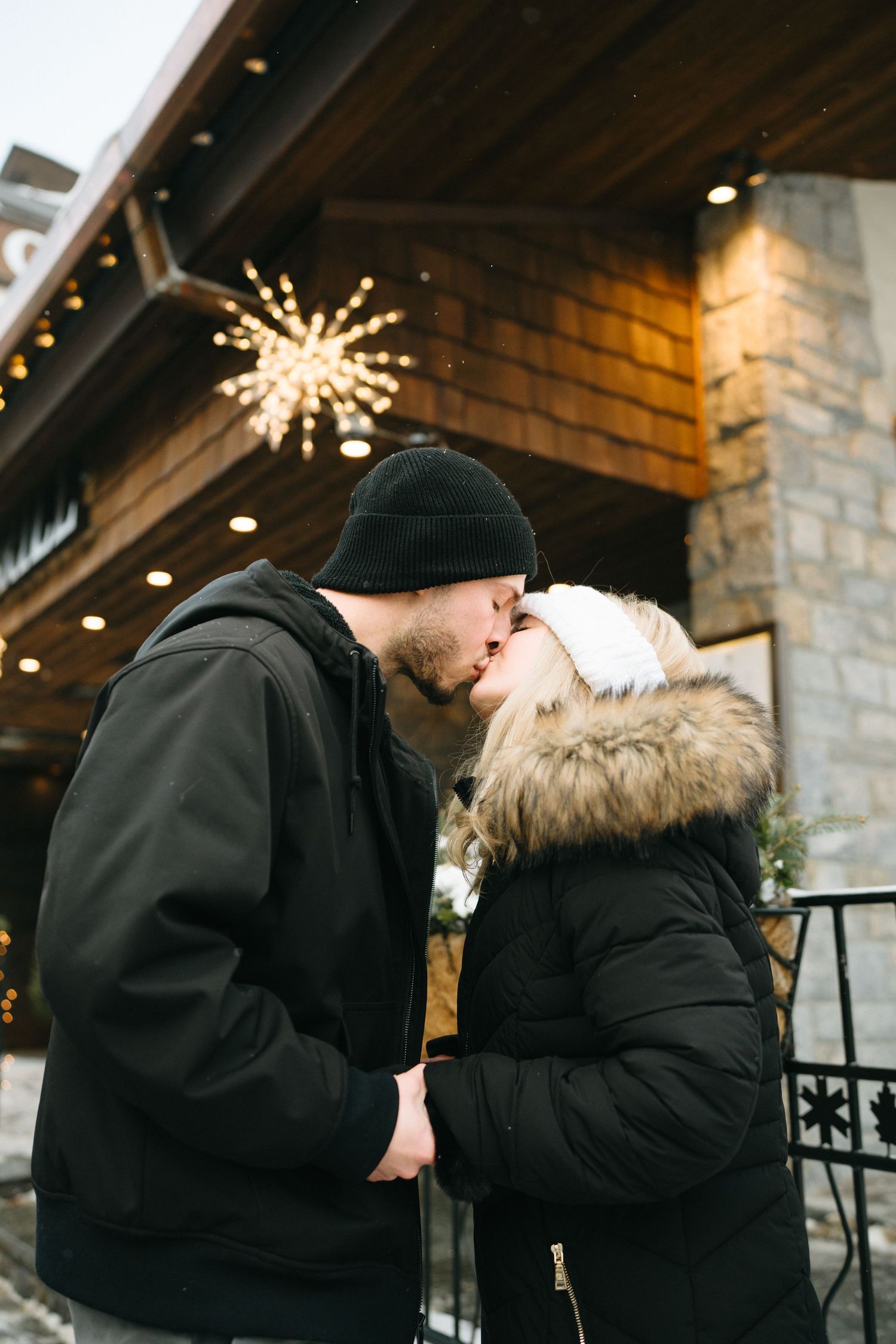 A romantic winter engagement photo sequence showing a couple kissing under glowing snowflake lights at night.