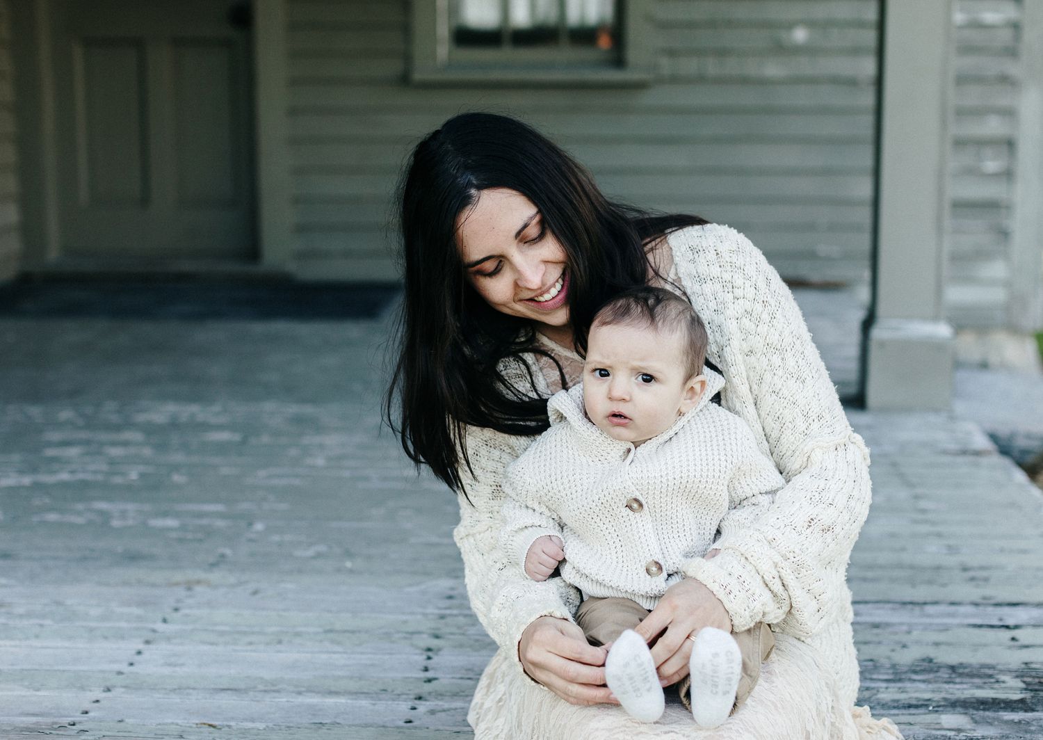 Mother wearing a lace cream dress and sweater holds five month son on lap wearing a cream sweater for motherhood photos on weathered porch