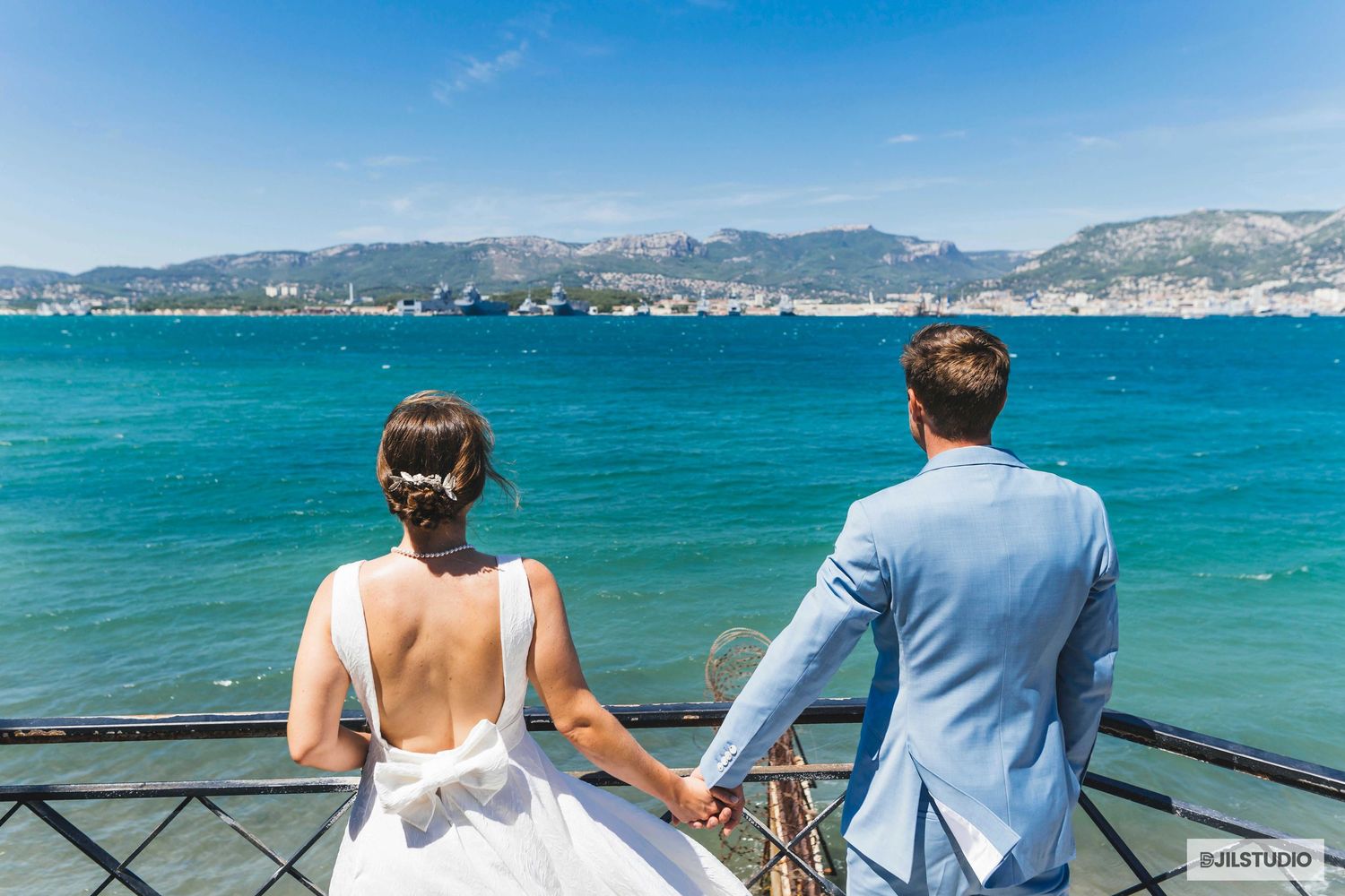 A couple in wedding attire stands at a railing overlooking turquoise Mediterranean waters and distant mountains.