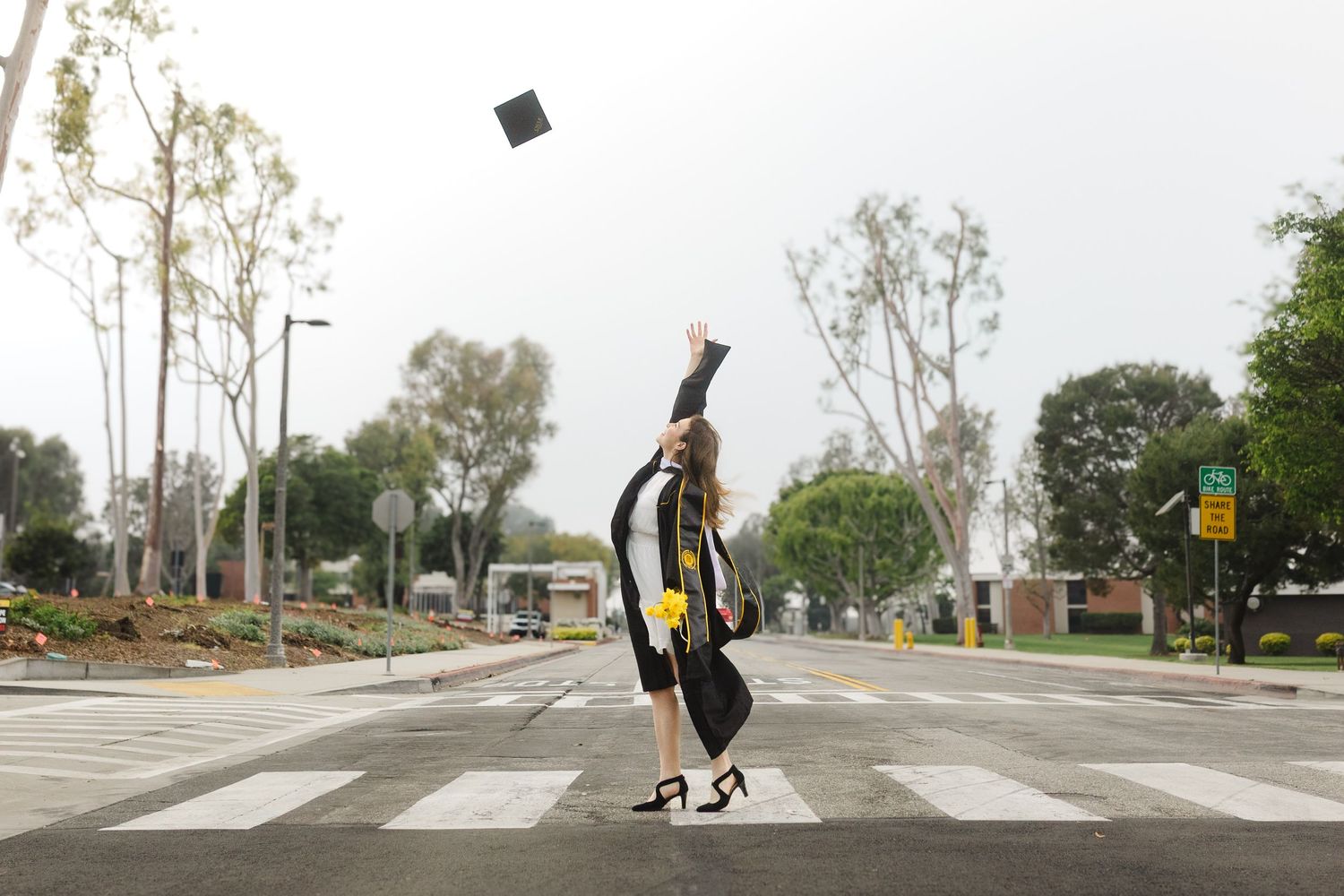 Graduate in cap and gown tosses mortarboard in air at crosswalk, celebrating academic achievement outdoors.
