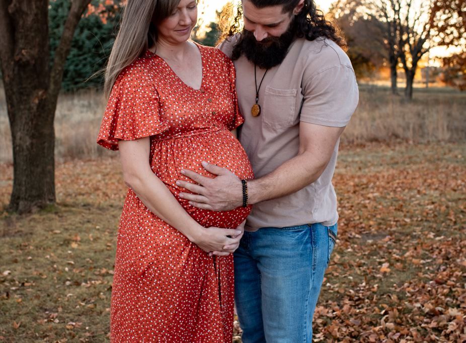 A couple in a red dress and casual attire embrace tenderly in a wooded area during golden hour sunset.