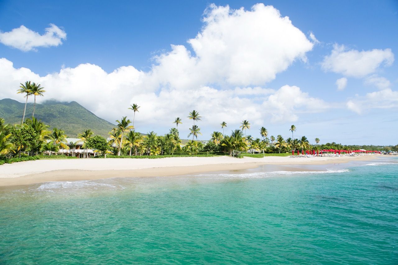 Tropical beach with turquoise water, white sand, palm trees, and lush green hills under a bright blue sky.