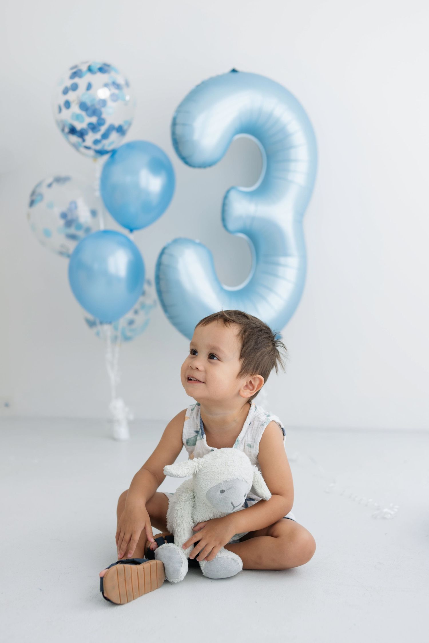 Toddler boy sitting on white floor holding stuffed animal toy with blue number 3 birthday balloons behind him.