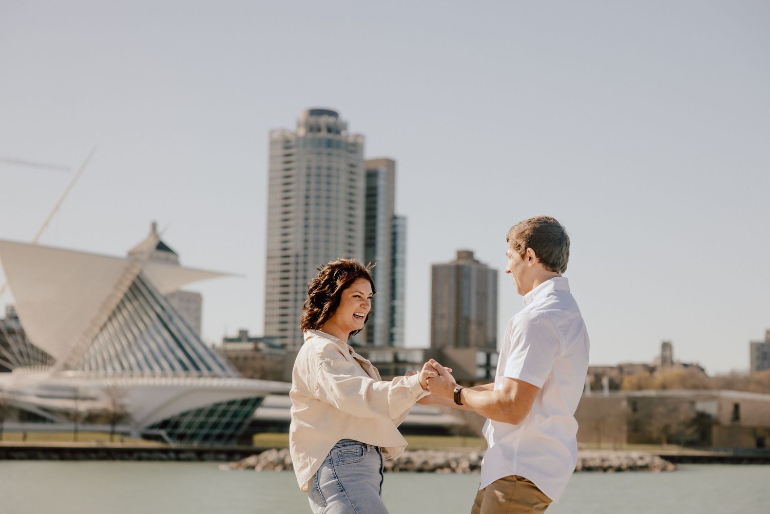 Couple dancing and holding hands outdoors near a waterfront with city skyline and modern architecture in background.