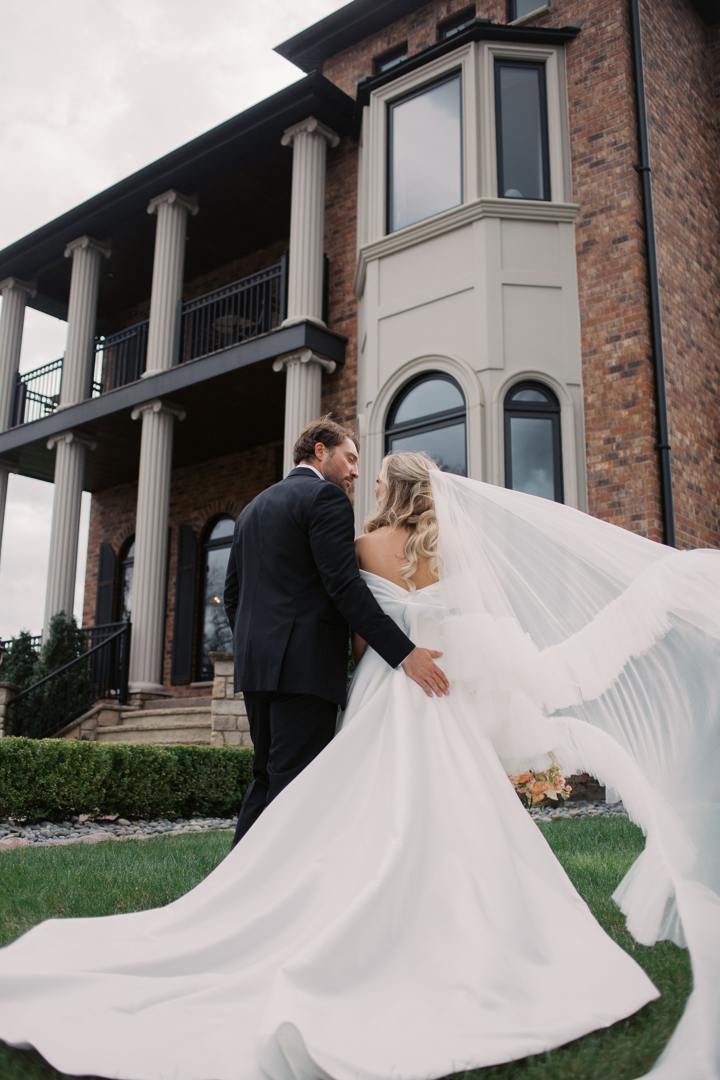 A bride and groom face away from the camera towards the Estate Washington Wedding venue. A cathedral veil blows in the wind.