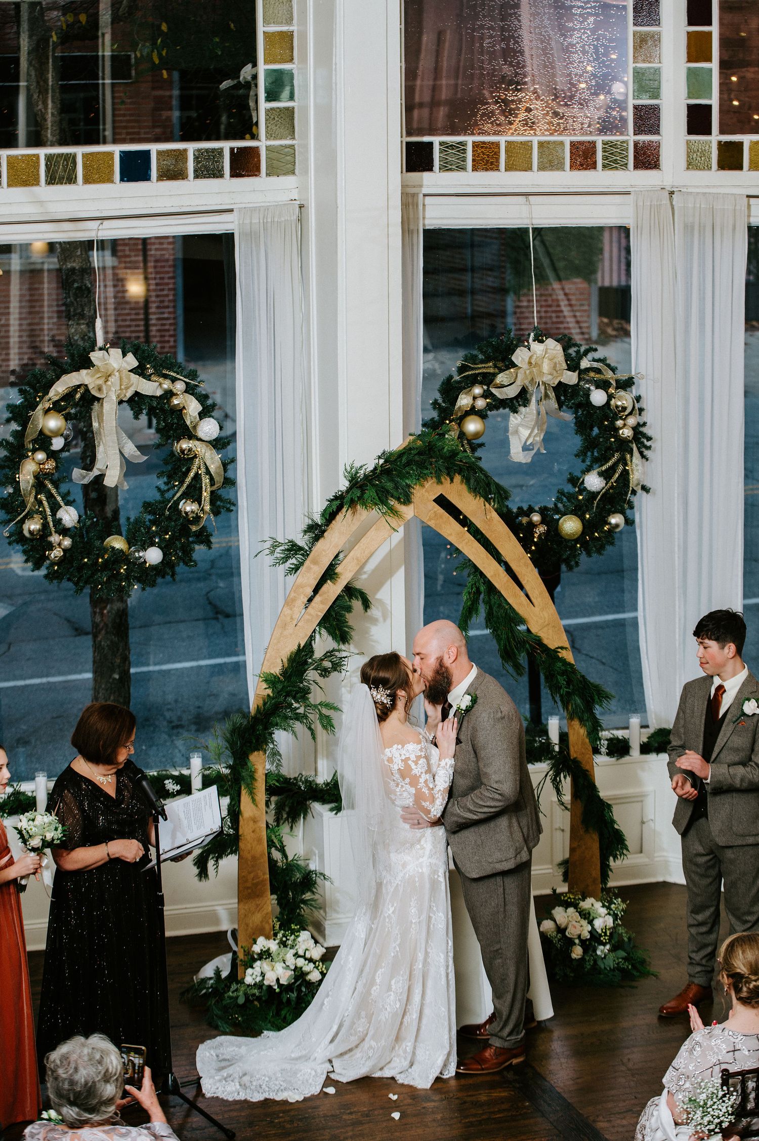 Wedding couple shares their first dance under wooden arches decorated with greenery and white flowers at an indoor reception.