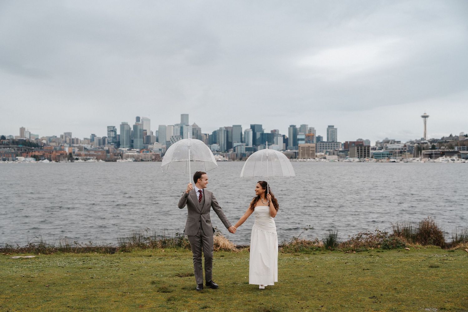 A couple in wedding attire poses on a grassy waterfront with the Seattle skyline visible in the background.