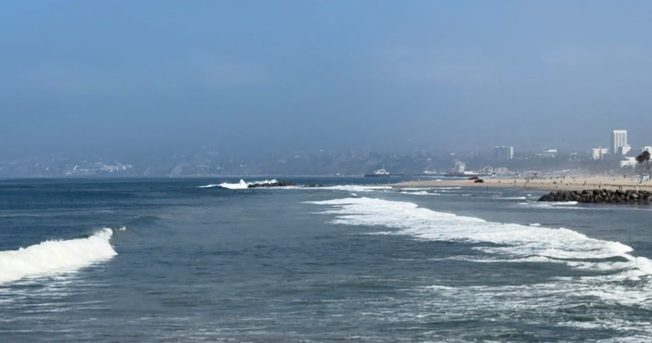 Waves rolling onto a sandy beach with a city skyline visible along the coastline under a hazy blue sky.
