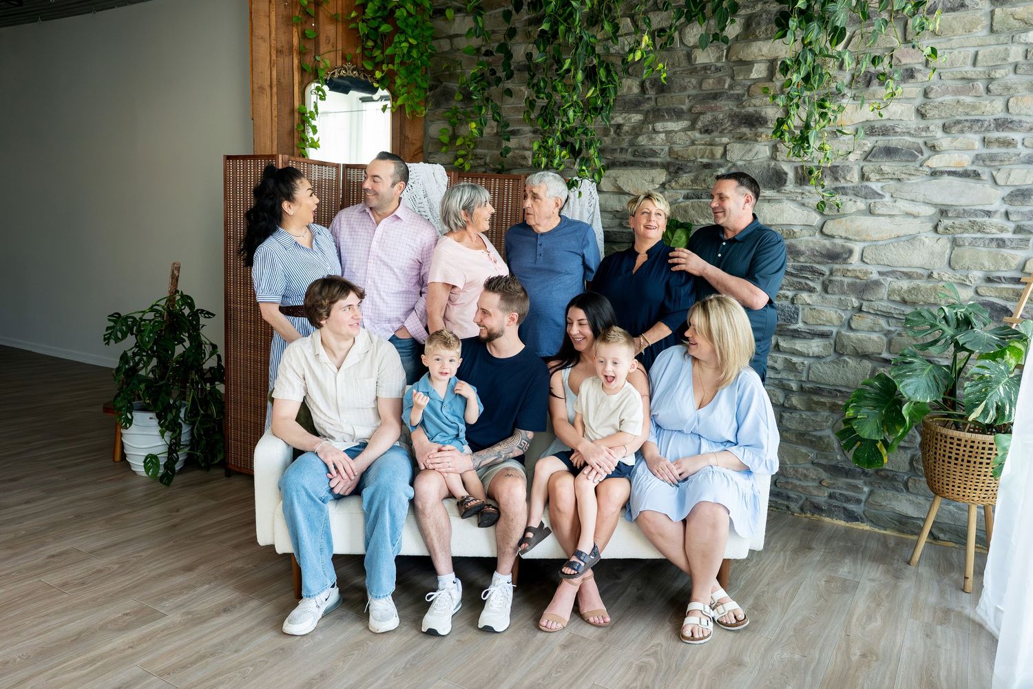 Large multi-generational family portrait gathered on white sofa with stone wall backdrop.