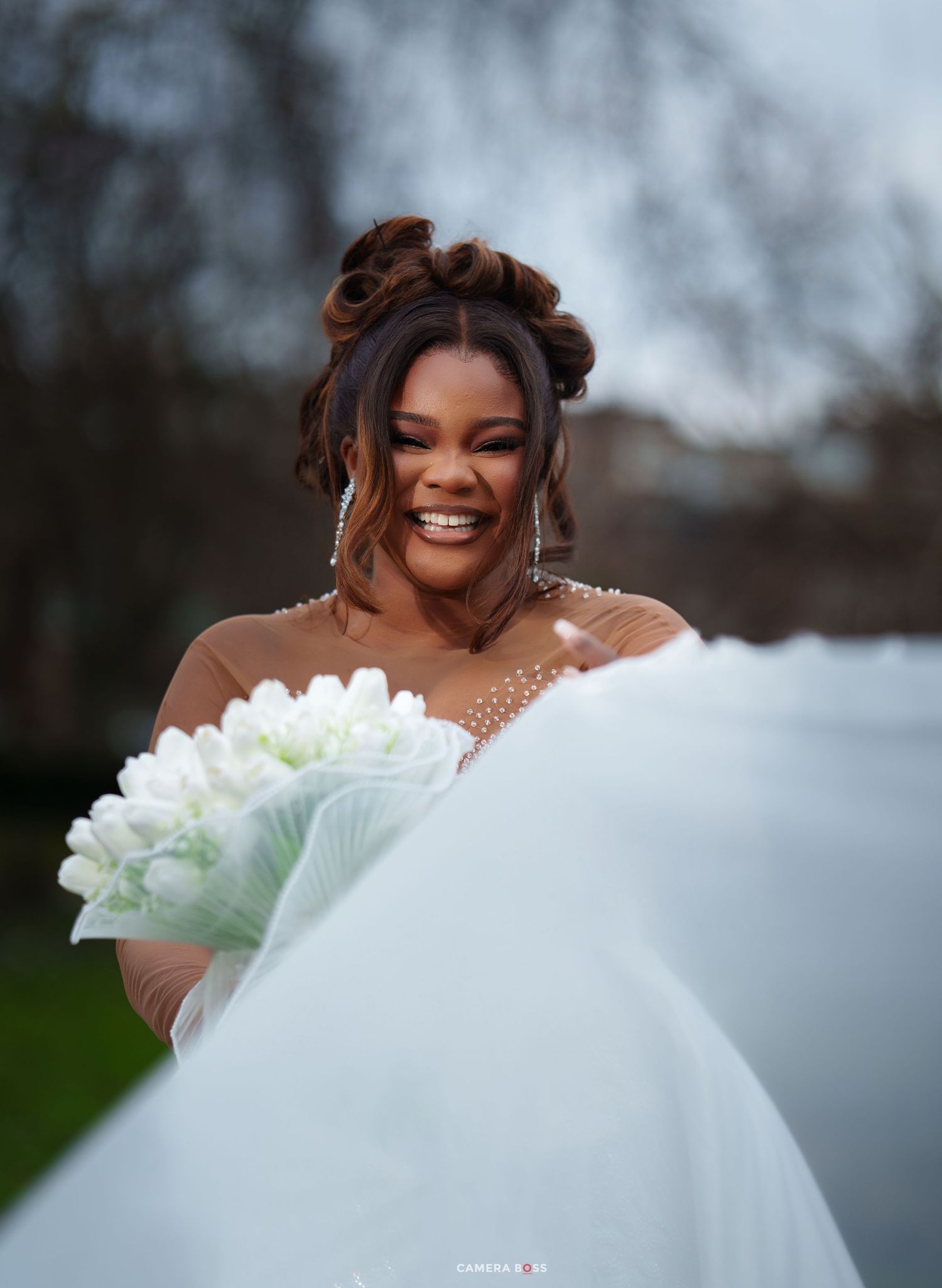 Smiling Black bride in white gown holds tulip bouquet outdoors, veil flowing, wearing elegant updo and drop earrings.