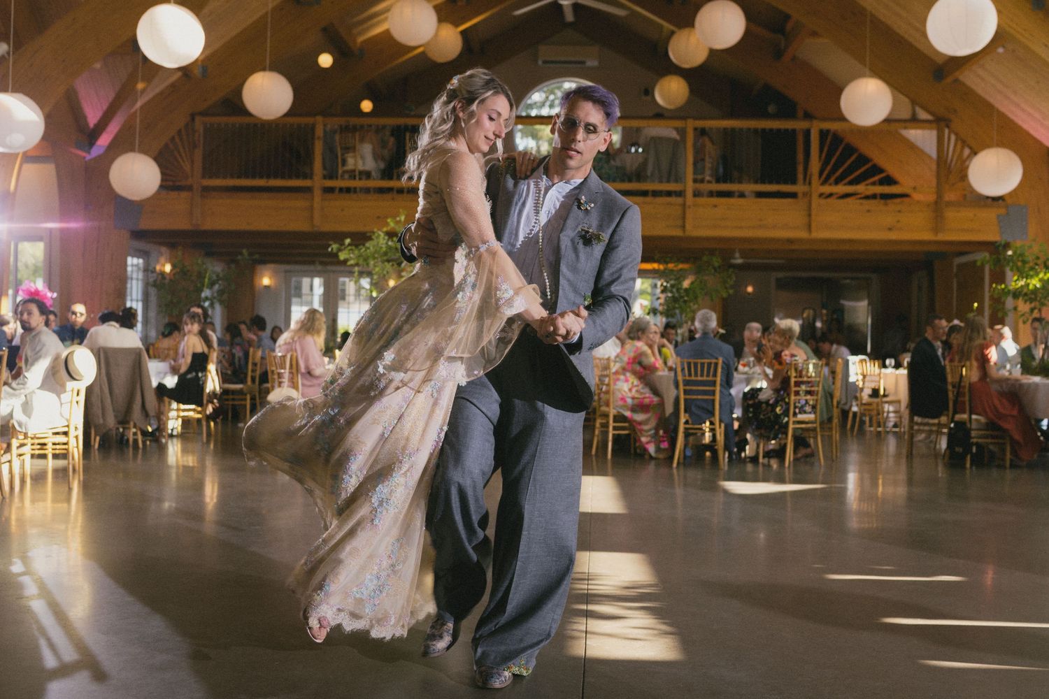 Bride and groom share their first dance at a rustic wedding reception with warm lighting and guests seated nearby.