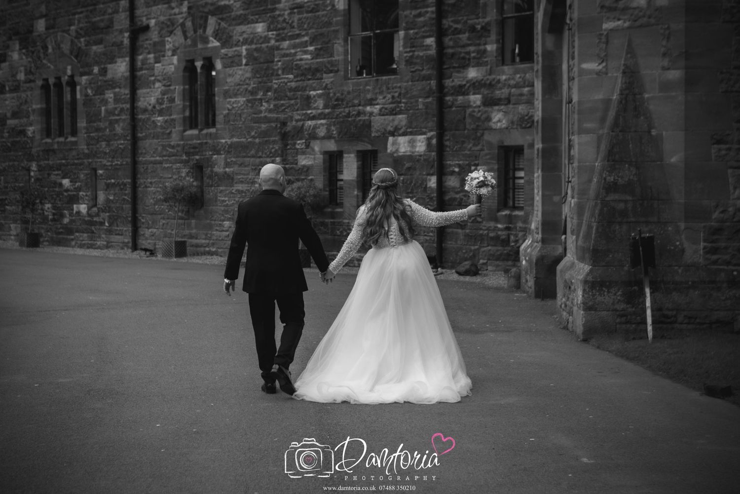 Large wedding party gathered in front of historic stone castle with Gothic arched windows and doorway on green lawn.