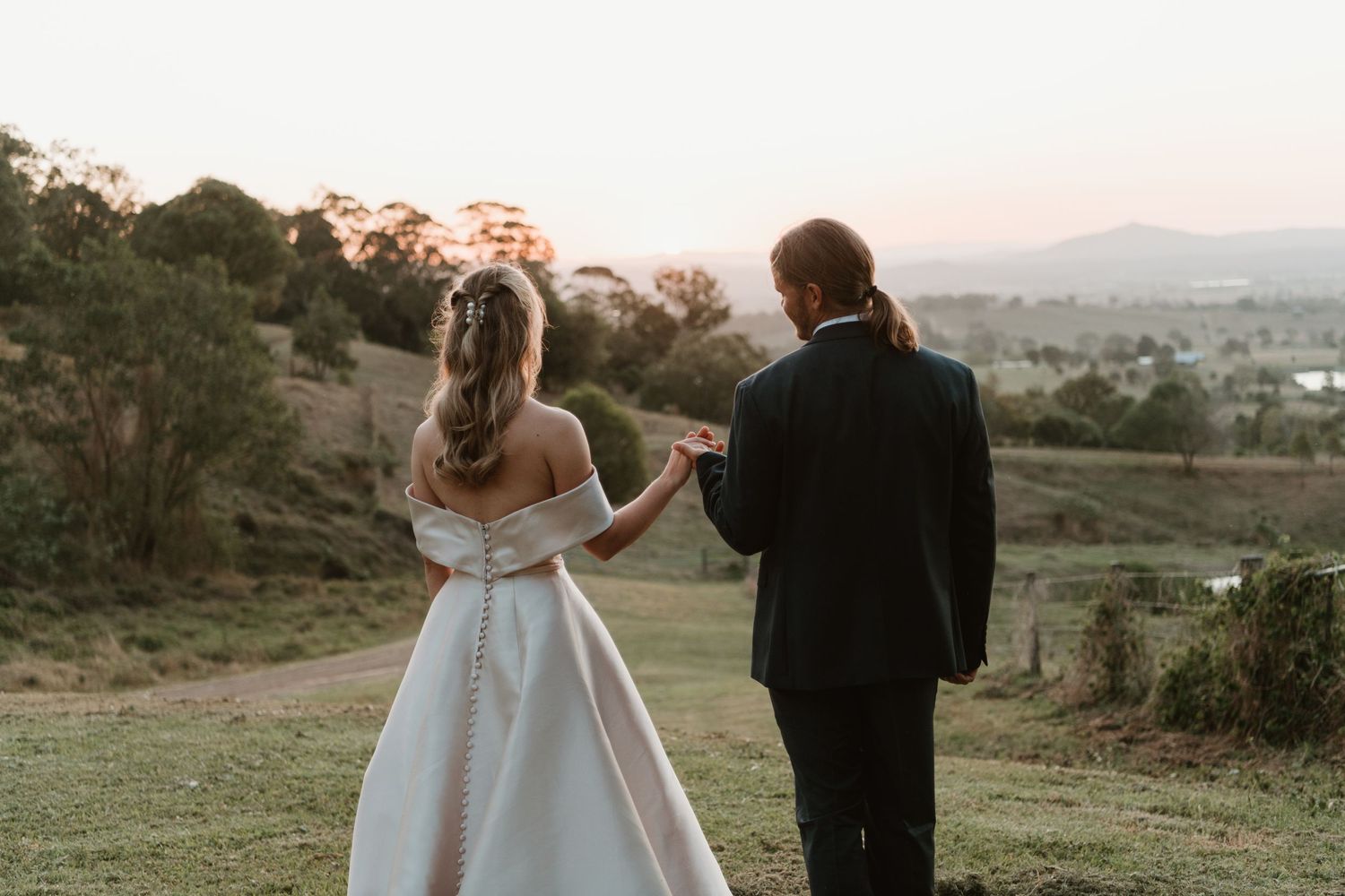 A bride and groom walk hand in hand at sunset overlooking a scenic green valley on their wedding day.
