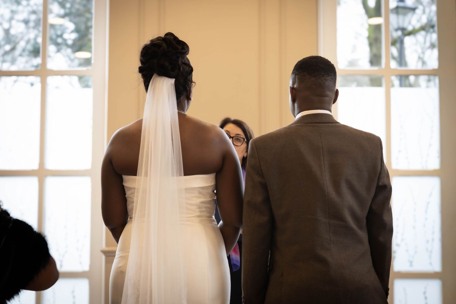 Black bride in white gown with veil stands beside groom in gray suit during indoor wedding ceremony.