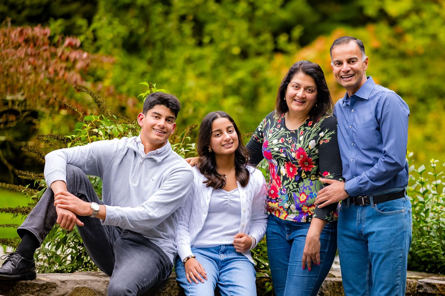 Family of four smiling outdoors in a lush green garden setting, posing together for a portrait photo.